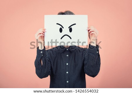 Businesswoman covering face using a white paper sheet with angry sad emoticon sketch, like fake mask for hiding the real emotion from society. Introvert female anonymity concept over pink wall. Royalty-Free Stock Photo #1426554032