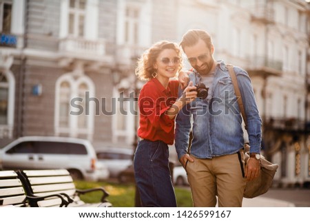 attractive smiling man and woman traveling together, stylish couple in love taking pictures on digital camera on romantic trip, sunny summer city, wearing shirt, sunglasses, travelers having fun