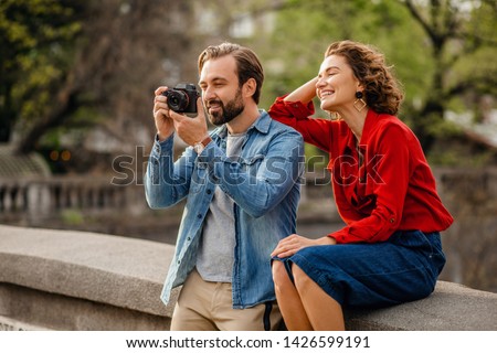 attractive happy smiling man and woman traveling together in Europe, stylish couple in love sitting in street taking photos on digital camera on romantic trip, sunny summer city, having fun
