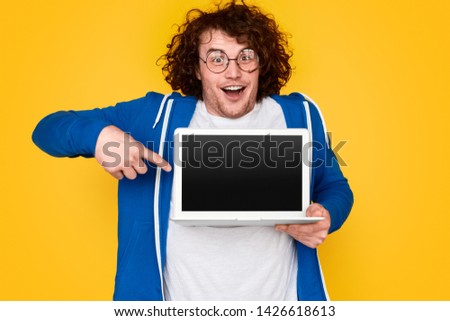Cheerful nerdy student with funny face pointing at modern laptop with empty screen against bright yellow background