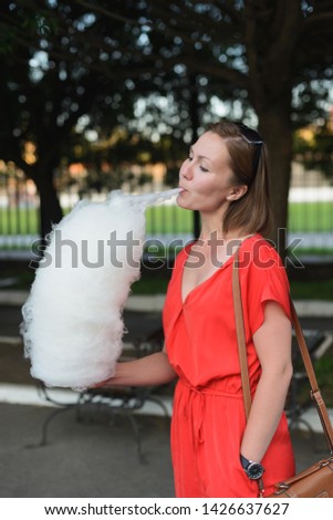 young adult caucasian woman in red dress eating cotton candy at public park, vertical lifestyle stock photo image