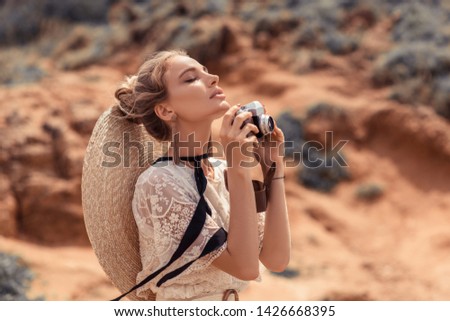 Fashionable image of amazing blonde woman taking photo with retro camera outdoor. Stylish holiday summer outfit. Boho accessories. 