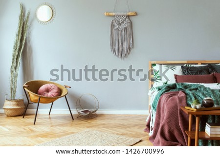 Stylish and luxury interior of bedroom with design furnitures, honey yellow armchair, gray macrame, gold mirror and elegant accessories. Beautiful bed sheets, blankets and pillows. Cozy home decor.