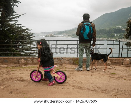Candid photo of daughter riding her little bicycle as father contemplates the shore from lookout as they discover the place on holidays.