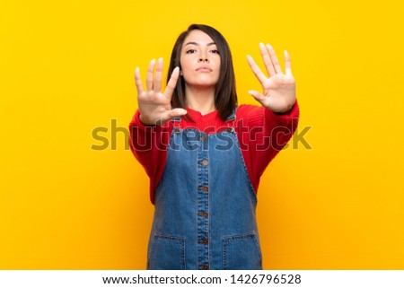 Young Mexican woman with overalls over yellow wall making stop gesture and disappointed