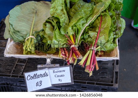 Freshly harvested bundles of collard greens and rainbow chard on display in a woven basket. Signage indicates vegetable type and price.