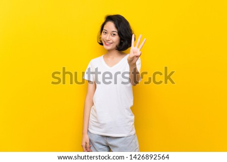 Asian young woman over isolated yellow wall happy and counting three with fingers