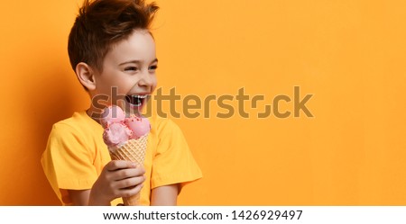 Baby boy kid in blank t-shirt with text space hold strawberry pink ice-cream in waffle cone happy laughing looking at the corner on yellow background