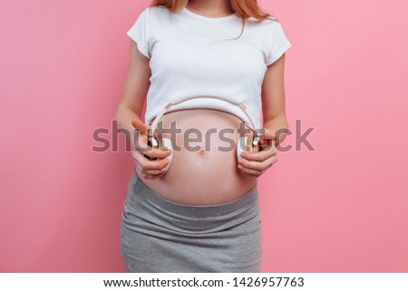 Attractive pregnant woman holding headphones on her belly over a pink background. The concept of pregnancy, motherhood, training and expectations