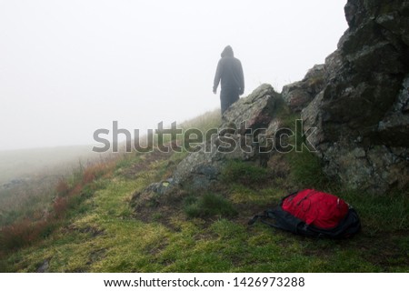 An abandoned rucksack on a bleak rocky moorland, with a hooded figure standing in the background on a foggy spooky day