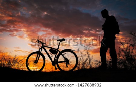 Silhouette of sports person cycling on the meadow on the beautiful sunset. Young man with camera and bicycle.