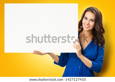 Happy smiling beautiful young woman showing blank sign board with copy space, isolated over yellow orange  background. Success in business concept. Brunette model in blue confident clothing at studio.