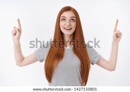 Amused dreamy thrilled joyful ginger girl blue eyes open mouth amazed look pointing index fingers up excited enjoy watching beautiful clouds fly sky standing daydreaming white background