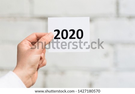 Business concept. White sheet with black 2020 text in man hand in front of white bricks wall, panorama, copy space
