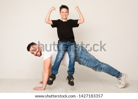 Family of strongmen. Dad doing push ups with son on back, cheerful boy showing biceps on camera Royalty-Free Stock Photo #1427183357