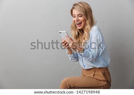 Photo of alluring blond businesswoman with long curly hair smiling while listening to music with smartphone and earphones isolated over gray background