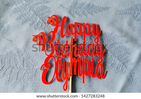 Beautiful red Happy birthday sign Olympia mounted on a wooden stick laid on a table with a tablecloth. He was pricked in a birthday party cake.