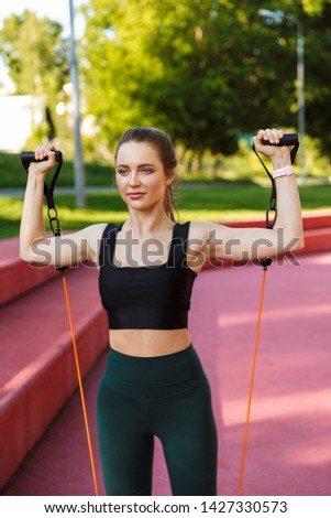 Photo of happy sporty woman 20s wearing tracksuit stretching her arms with expander fitness rope during morning workout in green park