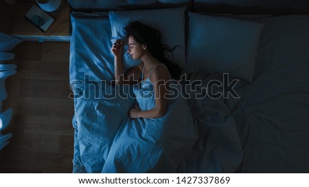 Top View of Beautiful Young Woman Sleeping Cozily on a Bed in His Bedroom at Night. Blue Nightly Colors with Cold Weak Lamppost Light Shining Through the Window. Royalty-Free Stock Photo #1427337869