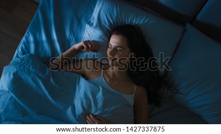 Top View of Beautiful Young Woman Sleeping Cozily on a Bed in His Bedroom at Night. Blue Nightly Colors with Cold Weak Lamppost Light Shining Through the Window. Royalty-Free Stock Photo #1427337875