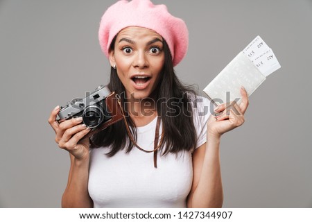 Photo closeup of surprised pretty tourist woman in beret wondering while holding retro camera and travel tickets isolated over gray background