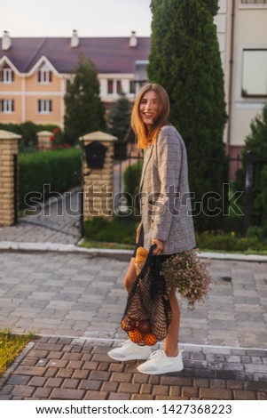 Beautiful girl in square jacket hold of flowers, string bag with fruits and baguette go to home. Romantic girl come to private house with trees. Photo from side and girl is turn around. Housewife.