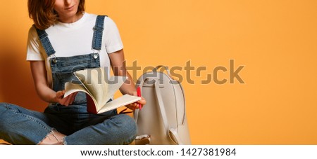 Cropped photo of a pensive girl in a white T-shirt and denim overalls, sitting on the floor with a kransny notebook and a pencil on a yellow background and writing something