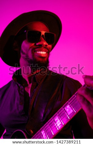 Young african-american musician playing the guitar like a rockstar on gradient purple-pink background in neon light. Concept of music, hobby. Joyful attractive guy improvising. Colorful portrait.