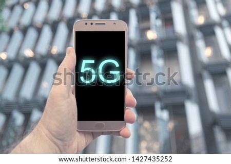 Hand holding a 5G compatible mobile smartphone against a modern building facade in Barelona. Empty copy space for Editor's text.