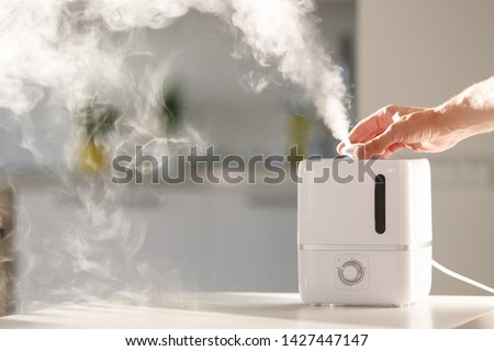 Hand turn on aroma oil diffuser on the table at home, steam from the air humidifier, free space. Ultrasonic technology, increase in air humidity indoors, comfortable living conditions, moisture.  Royalty-Free Stock Photo #1427447147