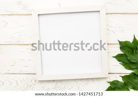 Summer flat lay with white photo frame and green leaves in corner on a white wooden background. Top view mockup with copy space for lettering