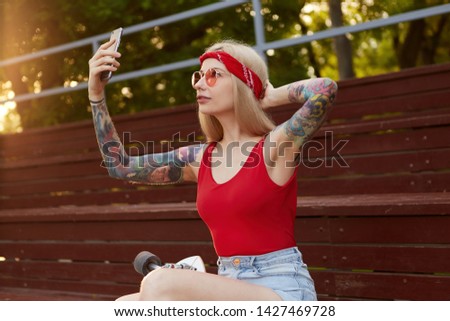 Portrait of young beautiful blonde woman with tattooed arms in a red T-shirt and denim shorts, with a knitted bandana on head, in red glasses, holding smartphone and makes selfie for her blog.