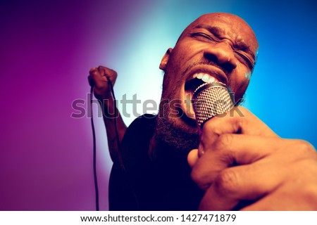 Young african-american jazz musician singing a song on gradient purple-blue background. Concept of music, hobby. Joyful attractive guy improvising, having a concert. Colorful retro portrait of singer.