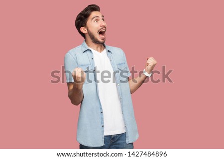Portrait of happy screaming handsome bearded young man in blue casual style shirt standing with suprised face and rejoicing his victory. indoor studio shot, isolated on pink background. Royalty-Free Stock Photo #1427484896