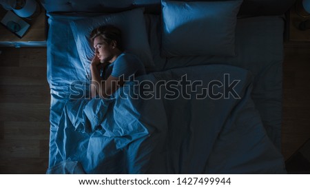 Top View of Handsome Young Man Sleeping Cozily on a Bed in His Bedroom at Night. Blue Nightly Colors with Cold Weak Lamppost Light Shining Through the Window. Royalty-Free Stock Photo #1427499944