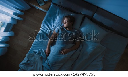 Top View of Handsome Young Man Sleeping Cozily on a Bed in His Bedroom at Night. Blue Nightly Colors with Cold Weak Lamppost Light Shining Through the Window. Royalty-Free Stock Photo #1427499953