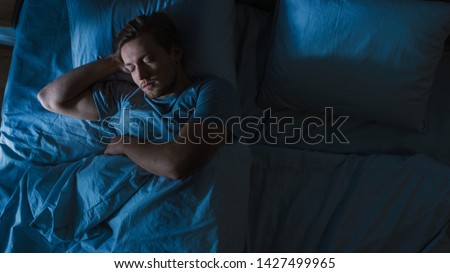 Top View of Handsome Young Man Sleeping Cozily on a Bed in His Bedroom at Night. Blue Nightly Colors with Cold Weak Lamppost Light Shining Through the Window. Royalty-Free Stock Photo #1427499965