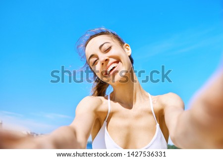 Portrait of beautiful happy woman taking a selfie on the camera phone with blue sky on background. Young smiling woman having fun and laughing against blue clear sky. Summertime and leisure concept.
