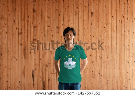 Man wearing Macau flag color of shirt and standing with crossed behind the back hands on the wooden wall background, green with a lotus and stylised Governor Nobre de Carvalho Bridge. 