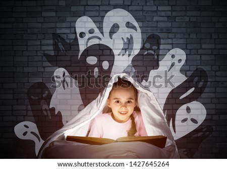 Happy little girl reading book in bed before going to sleep. Child with flashlight hiding under blanket. Portrait of smiling girl in pajamas and funny ghosts back on wall. Child reading fairy tales.