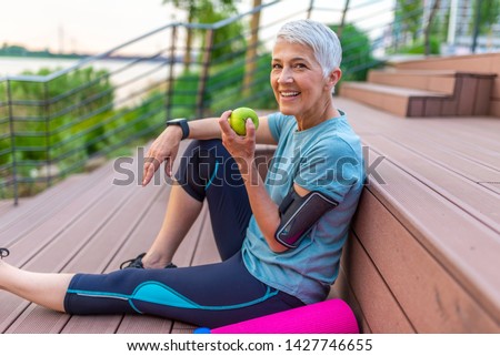 Sporty woman eating apple. Beautiful woman with gray hair in the early sixties relaxing after sport training. Healthy Age. Mature athletic woman eating an apple after sports training  Royalty-Free Stock Photo #1427746655