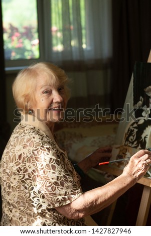 An elderly woman sits at the easel and paints in oils, sitting against window in sunlight.Silver age concept