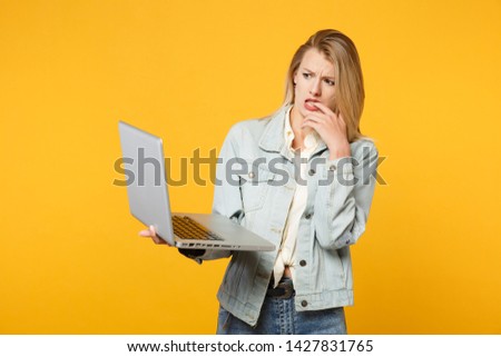 Portrait of concerned young woman in denim casual clothes gnawing nails holding, using laptop pc computer isolated on yellow orange background in studio. People lifestyle concept. Mock up copy space