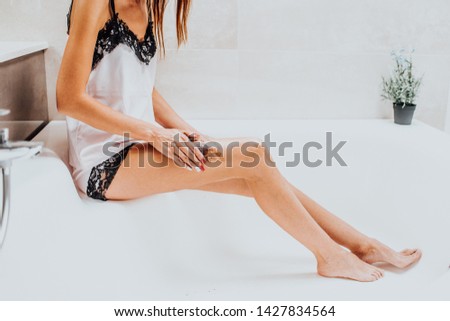 Slim young woman in a bright silk pajamas scrabs her leg sitting in a big beautiful bright bath. Skin care concept