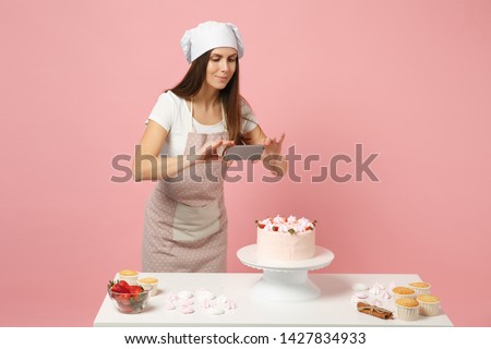 Chef cook confectioner or baker in apron white t-shirt, toque chefs hat cooking at table photographing cake for her blog isolated on pink pastel background in studio. Mock up copy space food concept