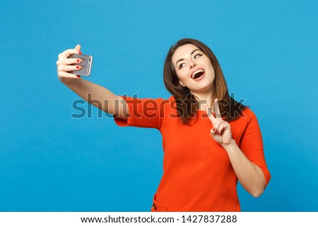 Beautiful brunette young woman wearing red orange dress doing selfie shot on mobile phone isolated over trendy blue background, studio portrait. People lifestyle fashion concept. Mock up copy space