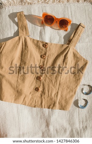 Brown linen top with wooden buttons and sunglasses with silver earrings lie on a beige carpet. Top view of fashion layout