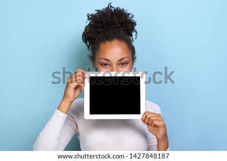 Mockup image of black empty blank screen of ipad in the female hand, peeking from behind tablet over blue background