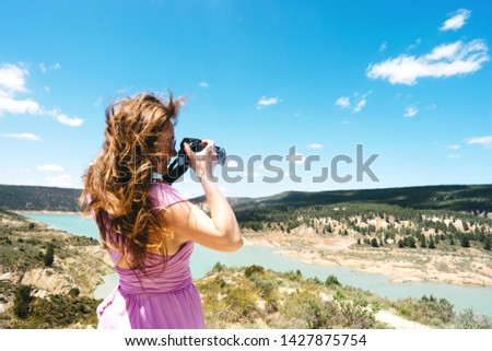 Long-haired female tourist in a pink dress with a camera in hand stands in the mountains against the backdrop of a beautiful lake. The concept of freedom, adventurism and travel