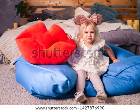 Photo of pretty blonde little girl in white dress holding big red lips,kiss-shaped soft pillow on modern background. Child with red lips shape cushion, Valentine day, love concept. 
